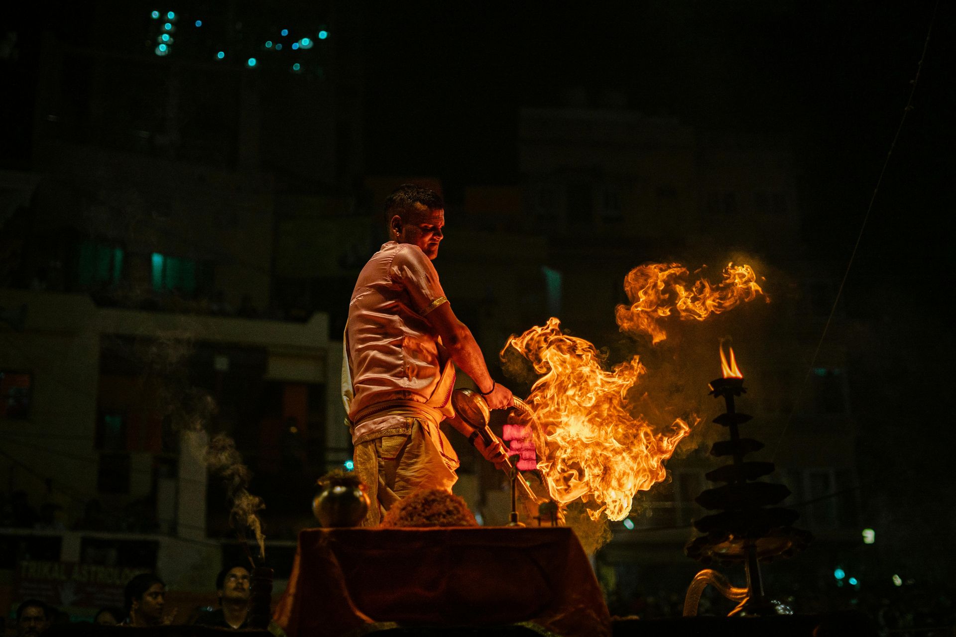 A Hindu priest performing the Ganga Aarti at night on Assi Ghat in Varanasi, holding a large brass lamp with flames rising high into the air. The fire illuminates his face, while the dark background is dotted with lights from nearby buildings.