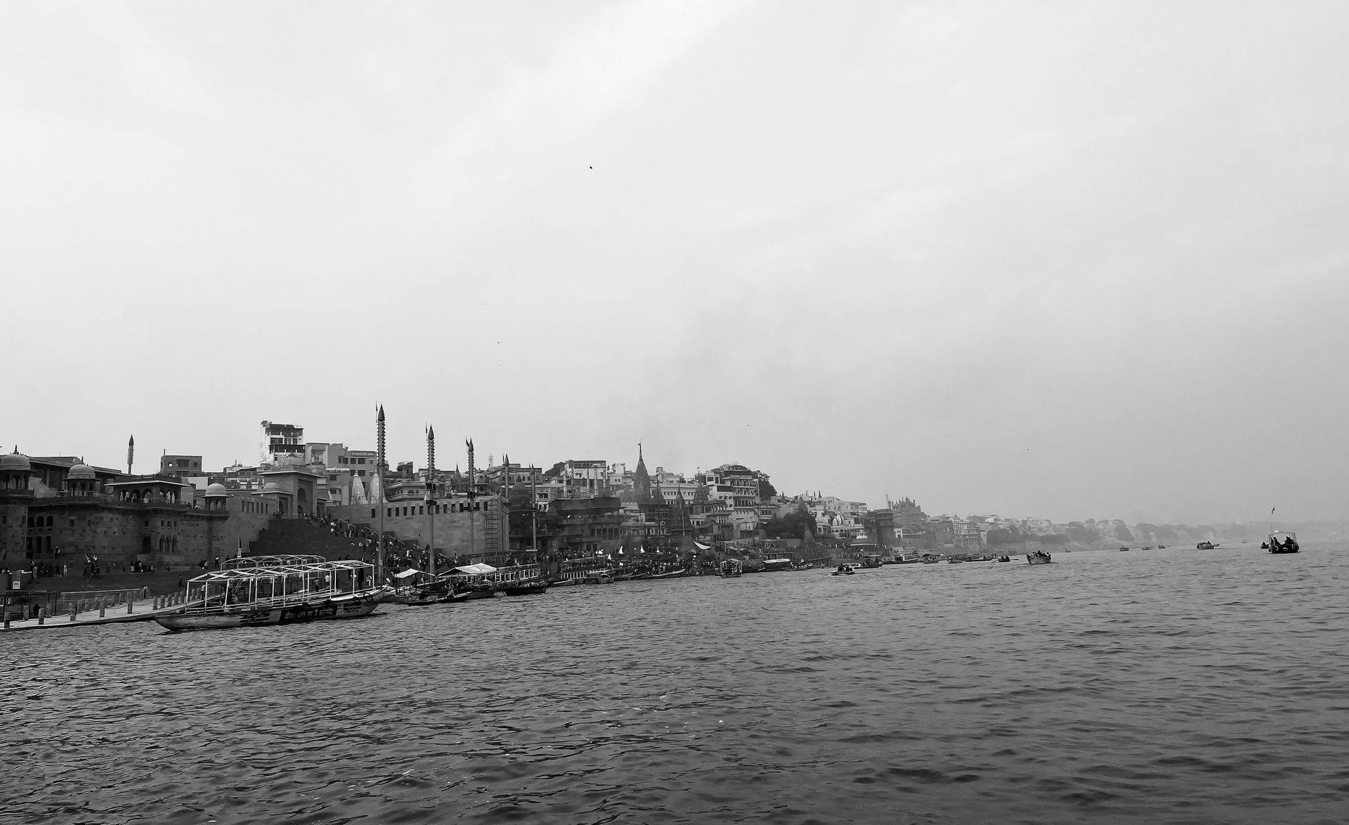 Varanasi’s ghats at dawn, viewed from a boat on the Ganges River. Historic temples and buildings line the riverbank, with boats floating nearby. A hazy sky and rising smoke from Manikarnika Ghat create a calm yet timeless atmosphere.