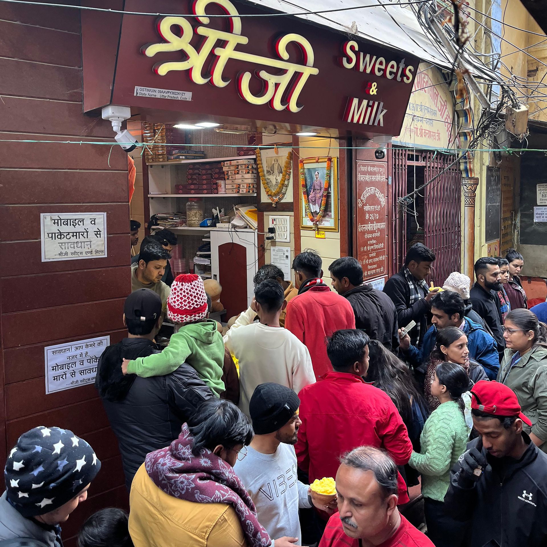  A crowded scene outside Shreeji Sweets & Milk in Varanasi, with people waiting in line to enjoy its famous malaiyo. The shop’s red signage is visible above the bustling crowd, capturing the city’s vibrant food culture.