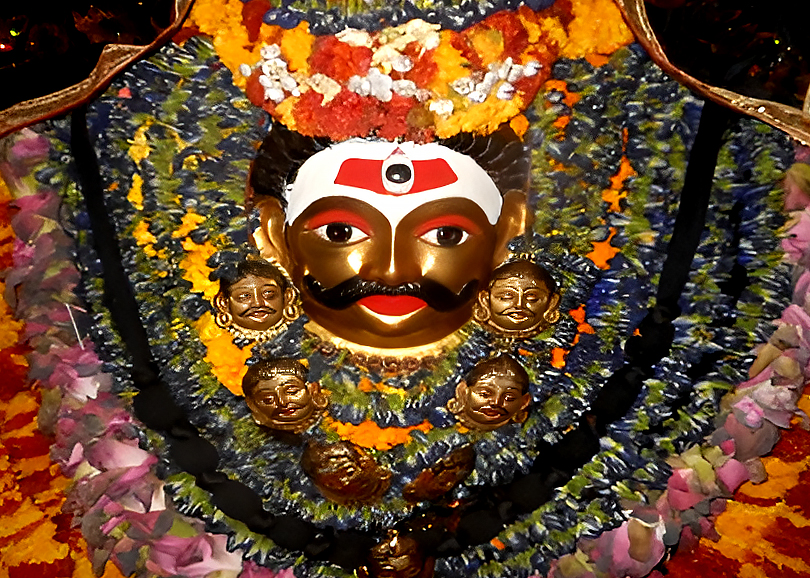 The idol of Kalbhairav at Kalbhairav Temple in Varanasi, adorned with a silver mask, a garland of skulls, and vibrant floral decorations. Worshipped as the guardian deity of the city, Kalbhairav is believed to hold the power over life and death in Varanasi.