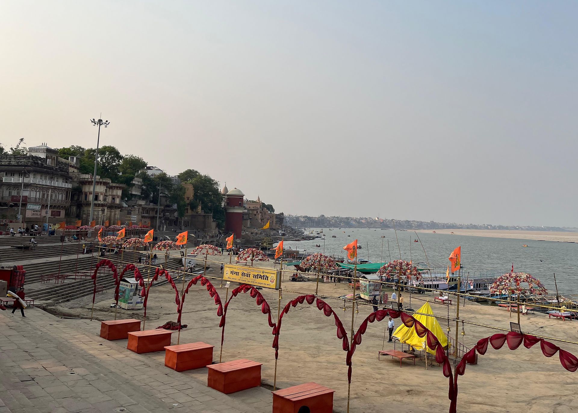 A serene view of Assi Ghat in Varanasi, with decorated archways, orange flags, and steps leading down to the Ganges River. The peaceful setting captures the quiet charm of the ghat as the sun sets, preparing for the evening aarti and spiritual gatherings.