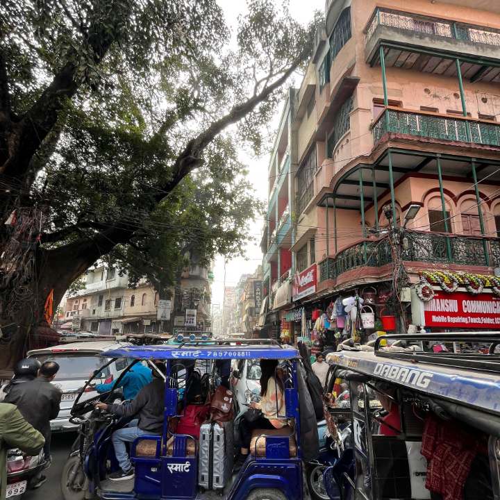 A bustling street in Varanasi filled with auto-rickshaws, pedestrians, and market shops. The crowded lanes, shaded by an old tree, capture the city’s unchanging rhythm as travelers depart.